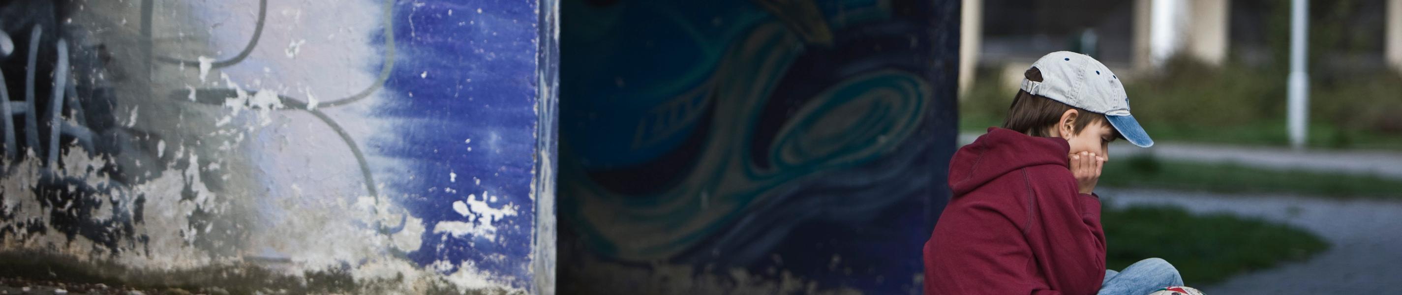 "ZDF.reportage: Alltag Kinderarmut -  Kein Geld - keine Chance?": Ein Junge seitlich zur Kamera, am rechten Bildrand mit Jeans, einer Kappe und einem roten Sweater. Er sitzt alleine mit seinem Fußball in einem ärmlichen Wohnviertel.