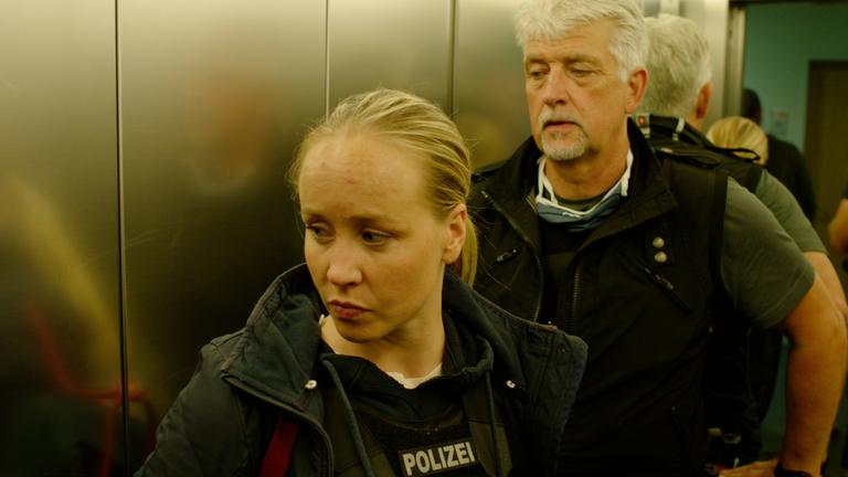 "37°: Auf der Spur der Täter - Delikt Kinderpornografie": Eine junge blonde Frau und ein weißhaariger Mann in Polizeiweste stehen im Fahrstuhl vor der Tür.