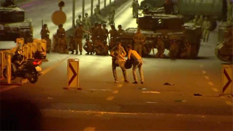 "Kampf auf der Bosporus-Brücke - Die Türkei und der gescheiterte Putschversuch": Zwei junge Männer tragen einen Verletzten auf der Bosporus-Brücke weg, im Hintergrund stehen zahlreiche Soldaten mit Maschinengewehren vor einem Panzer.
