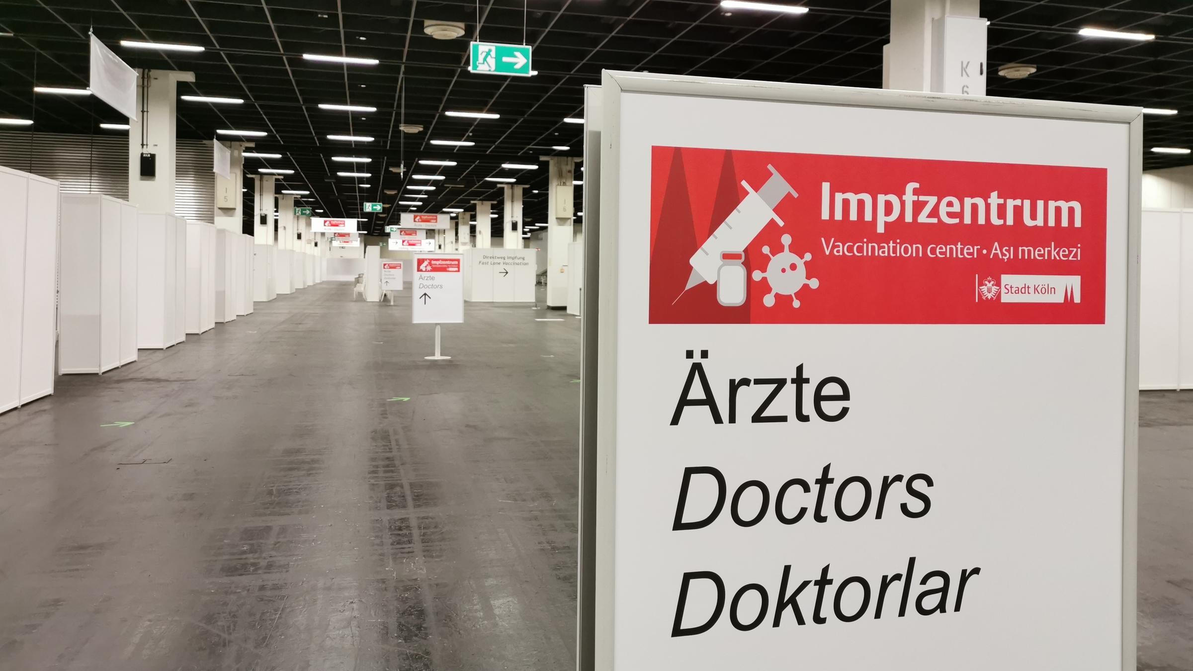 "ZDF.reportage: Impfen bitte! - Der harte Kampf gegen Corona": Eine menschenleere Messehalle, am linken Bildrand mehrere weiße Stände. Im Vordergrund ein Wegweiser mit der mehrsprachigen Aufschrift "Impfzentrum" und "Ärzte"