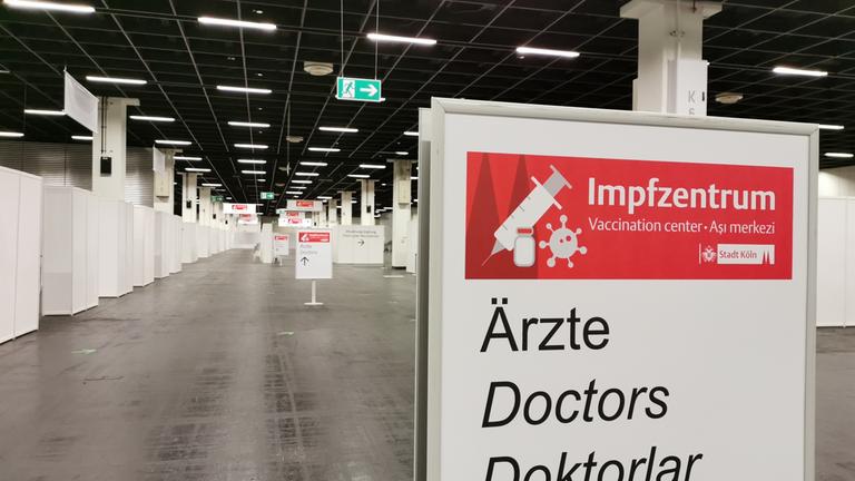 "ZDF.reportage: Impfen bitte! - Der harte Kampf gegen Corona": Eine menschenleere Messehalle, am linken Bildrand mehrere weiße Stände. Im Vordergrund ein Wegweiser mit der mehrsprachigen Aufschrift "Impfzentrum" und "Ärzte"