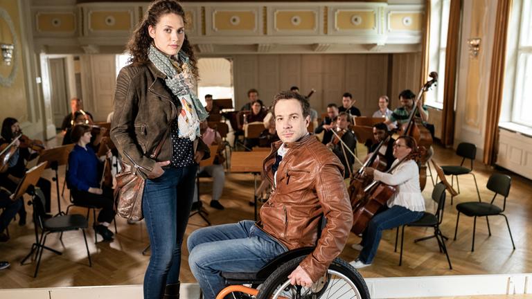 "Die Toten von Salzburg - Schwanengesang": Irene Russmeyer (Fanny Krausz) steht Peter Palfinger (Florian Teichtmeister) gegenüber, der in einem Rollstuhl sitzt. Hinter den beiden befindet sich ein Orchester mit mehreren Menschen.