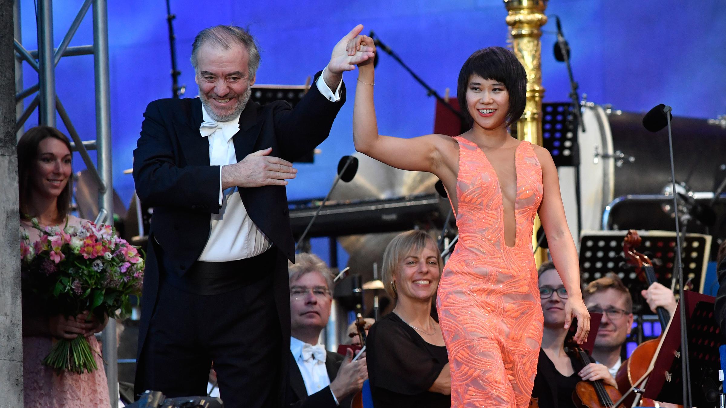 "Sommernachtsmusik - Open Air aus München": Der Chefdirigent der Münchner Philharmoniker, Valery Gergiev, zusammen mit der Pianistin Yuja Wang vor dem Orchester. Er im Frack, sie in einem orangefarbenen Kleid. Er hebt ihre Hand, das Orchester im Hintergrund applaudiert.