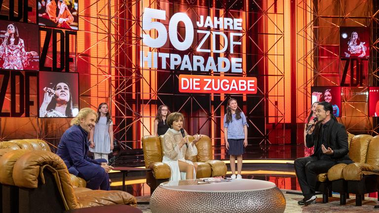 "50 Jahre ZDF-Hitparade - Die Zugabe": Paola Felix und Bülent Ceylan singen gemeinsam. Neben ihnen ist Thomas Gottschalk und lauscht ihnen freudig. Alle sitzen auf einem Sessel. Im Hintergrund sind Kinder zu sehen sowie das Hitparaden-Logo.