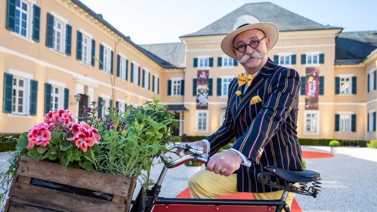 "Bares für Rares - Deutschlands größte Trödelshow": Moderator Horst Lichter steht an ein Fahrrad gelehnt, in dessen Rahmen das Sendungslogo zu sehen ist und lächelt in die Kamera. Im Hintergrund ist Schloss Johannisberg im Rheingau zu sehen.