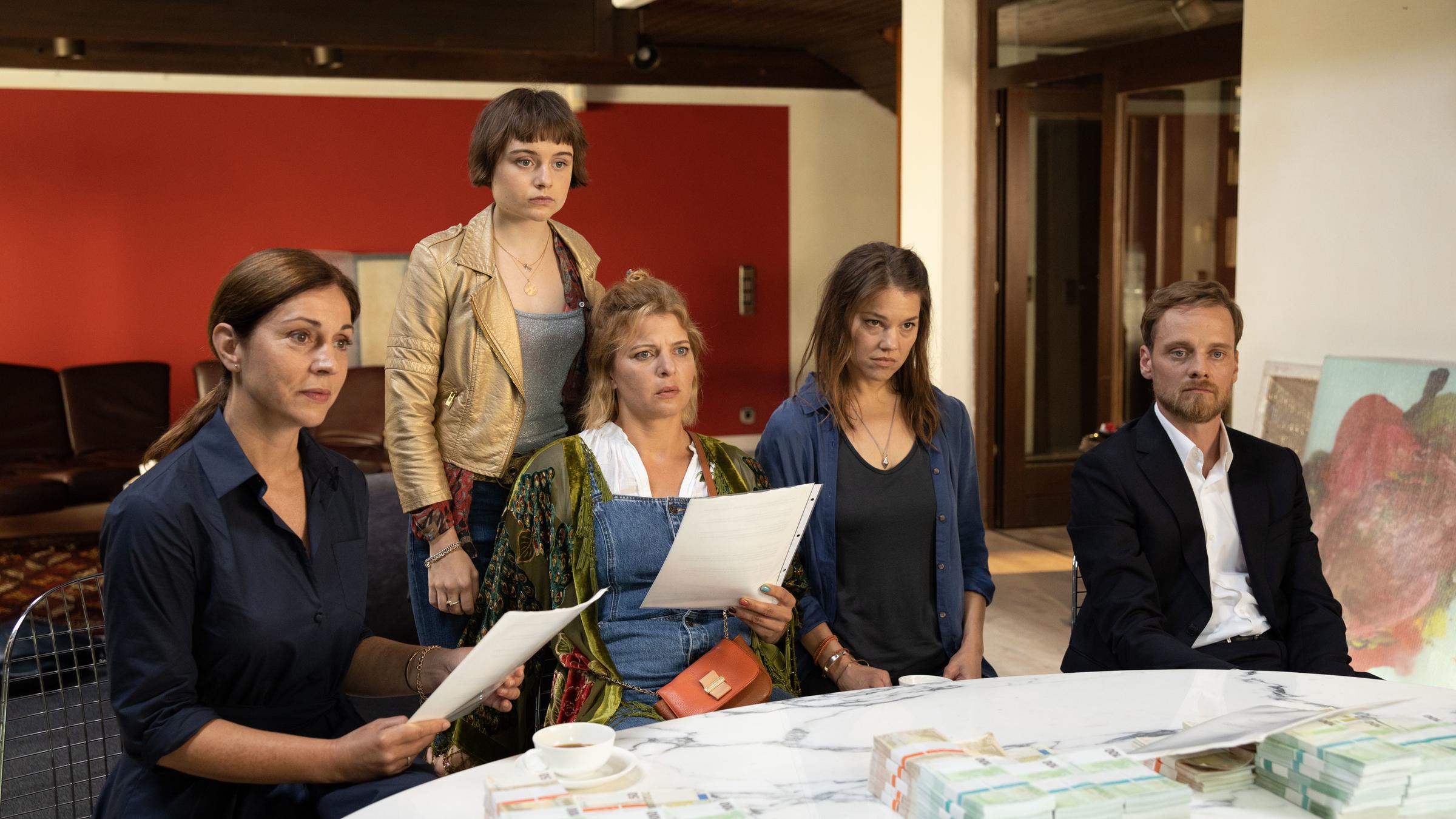 "Mutter kündigt": Drei Frauen und ein Mann sitzen an einem Tisch, auf dem sich Geldstapel türmen. Es sind die erwachsenen Geschwister Rita (Ulrike C. Tscharre), Doro (Jördis Triebel), deren Frau Hanna (Britta Hammelstein) und Phillipp (Stefan Konarske). In den Händen halten sie einen Vertrag. Hinter ihnen steht Joe (Lena Urzendowsky), Doros 18-jährige Tochter.