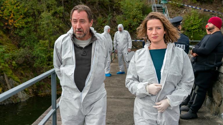 "Theresa Wolff": Theresa Wolff und Robert Brückner laufen über eine Brücke. Beide tragen Schutzanzüge.