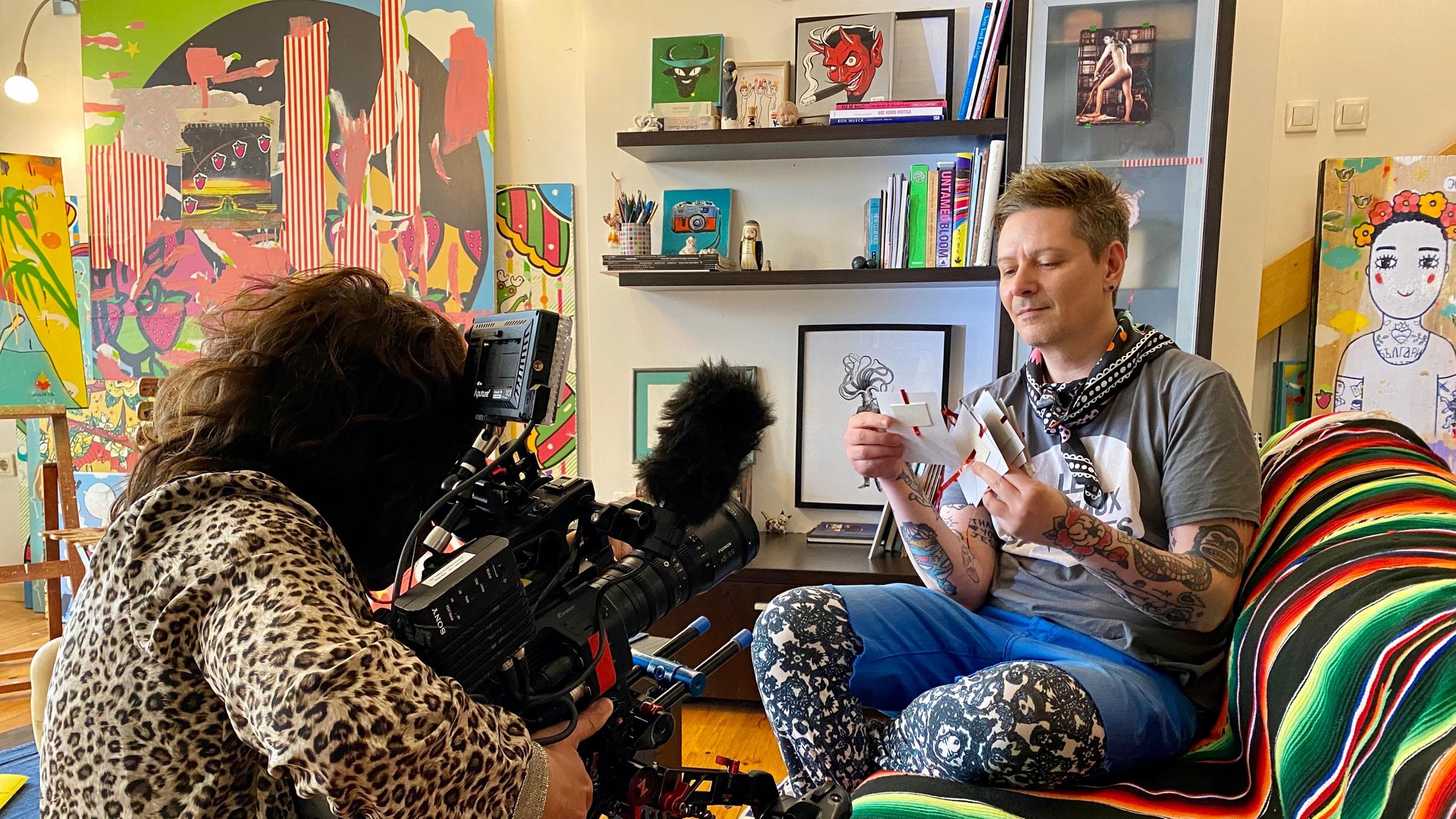 "Queer Balkan": In einem Wohnzimmer sitzt ein Mann auf dem Sofa, er hält ein Bündel Briefe in der Hand. Er hat kurze hellbraune Haare. Er trägt ein graues T-Shirt, eine blaue kurze Hose und darunter eine schwarz-weiß gemusterte Leggins. Seine Unterarme sind vollständig tätowiert. Links vor ihm kniet eine Kamerafrau und filmt ihn. Sie trägt ein Oberteil mit Leoparden-Print. Im Hintergrund ein Bücherregal und viele bunte Gemälde, die an die Wand gelehnt sind, hinten links eines auf einer Staffelei.
