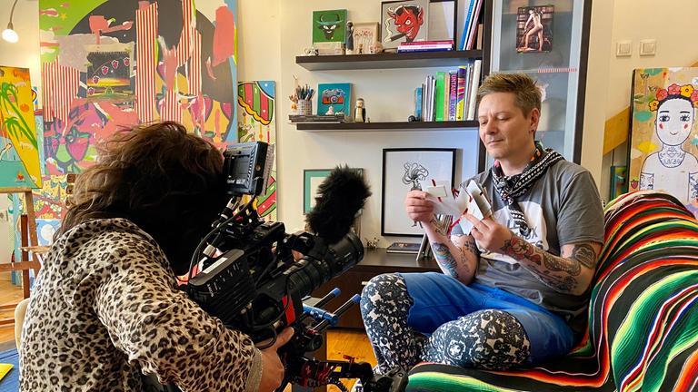 "Queer Balkan": In einem Wohnzimmer sitzt ein Mann auf dem Sofa, er hält ein Bündel Briefe in der Hand. Er hat kurze hellbraune Haare. Er trägt ein graues T-Shirt, eine blaue kurze Hose und darunter eine schwarz-weiß gemusterte Leggins. Seine Unterarme sind vollständig tätowiert. Links vor ihm kniet eine Kamerafrau und filmt ihn. Sie trägt ein Oberteil mit Leoparden-Print. Im Hintergrund ein Bücherregal und viele bunte Gemälde, die an die Wand gelehnt sind, hinten links eines auf einer Staffelei.