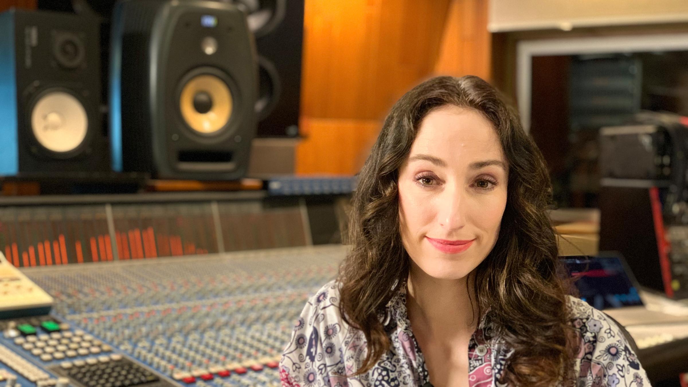 "Pop Around - The Video Stars": Eine Frau mit langen dunklen Locken sitzt in einem gemusterten Kleid vor einem großen Mischpult, im Hintergrund stehen zwei Boxen.