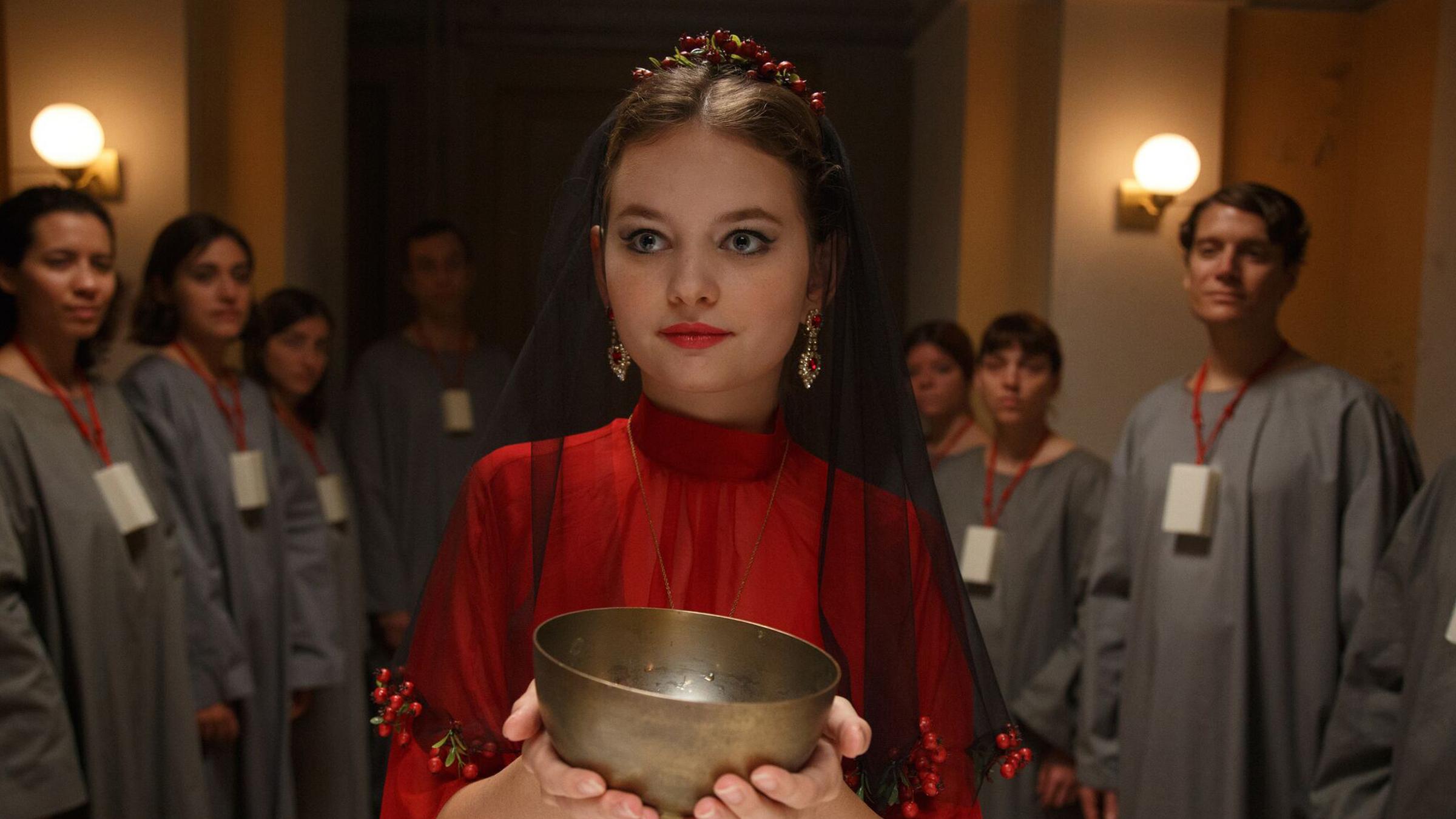 "A Pure Place": Irina (Greta Bohacek), eine junge Frau in rotem Gewand,  hält in einem abgedunkelten Raum einen Kelch in der Hand. Um sie herum stehen mit bewundernden Blicken weitere Menschen in grauen Gewändern, die ein Stück Seife um den Hals hängen haben.