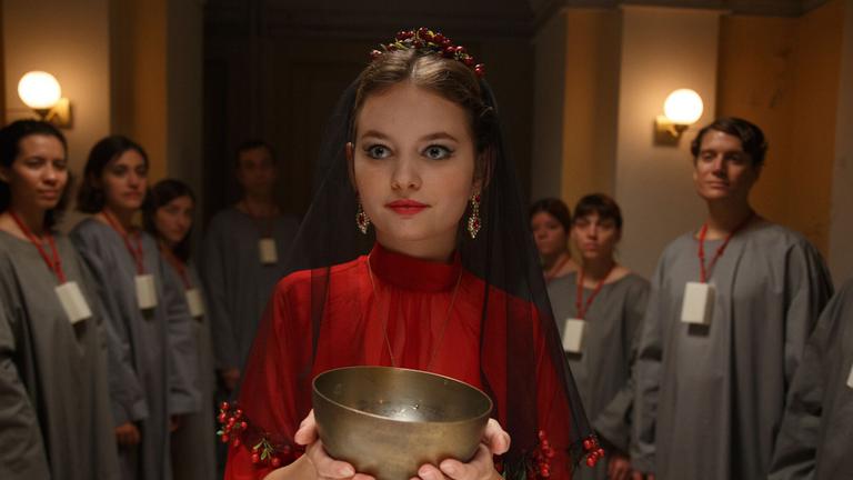 "A Pure Place": Irina (Greta Bohacek), eine junge Frau in rotem Gewand,  hält in einem abgedunkelten Raum einen Kelch in der Hand. Um sie herum stehen mit bewundernden Blicken weitere Menschen in grauen Gewändern, die ein Stück Seife um den Hals hängen haben.