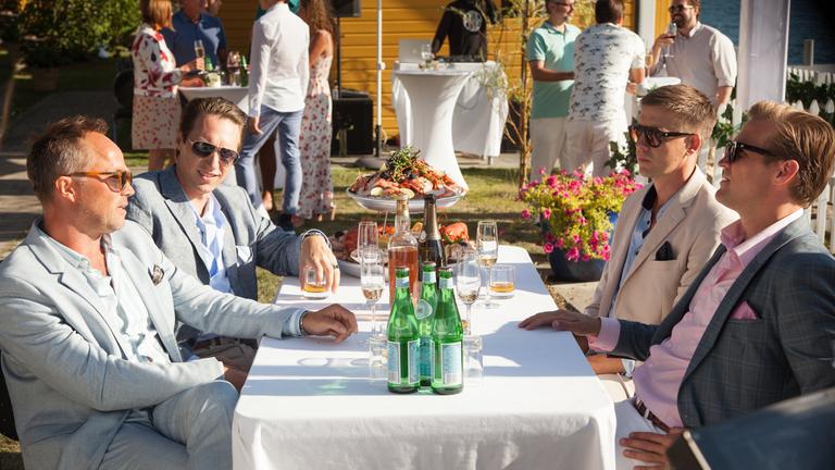 "Exit - Party": Adam Veile (Simon J. Berger) und Jeppe Schøitt (Jon Øigarden) sitzen Henrik Kranz (Tobias Santelmann) und William Bergvik (Pål Sverre Hagen) am gedeckte Tisch gegenüber und unterhalten sich. Alle tragen eine Sonnenbrille und Anzüge.