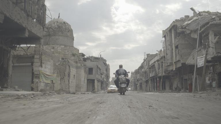 "Irak - Zerstörung eines Landes - Die Ölnation": Jemand fährt auf einem Motorrad durch eine völlig zerstörte  Stadt.