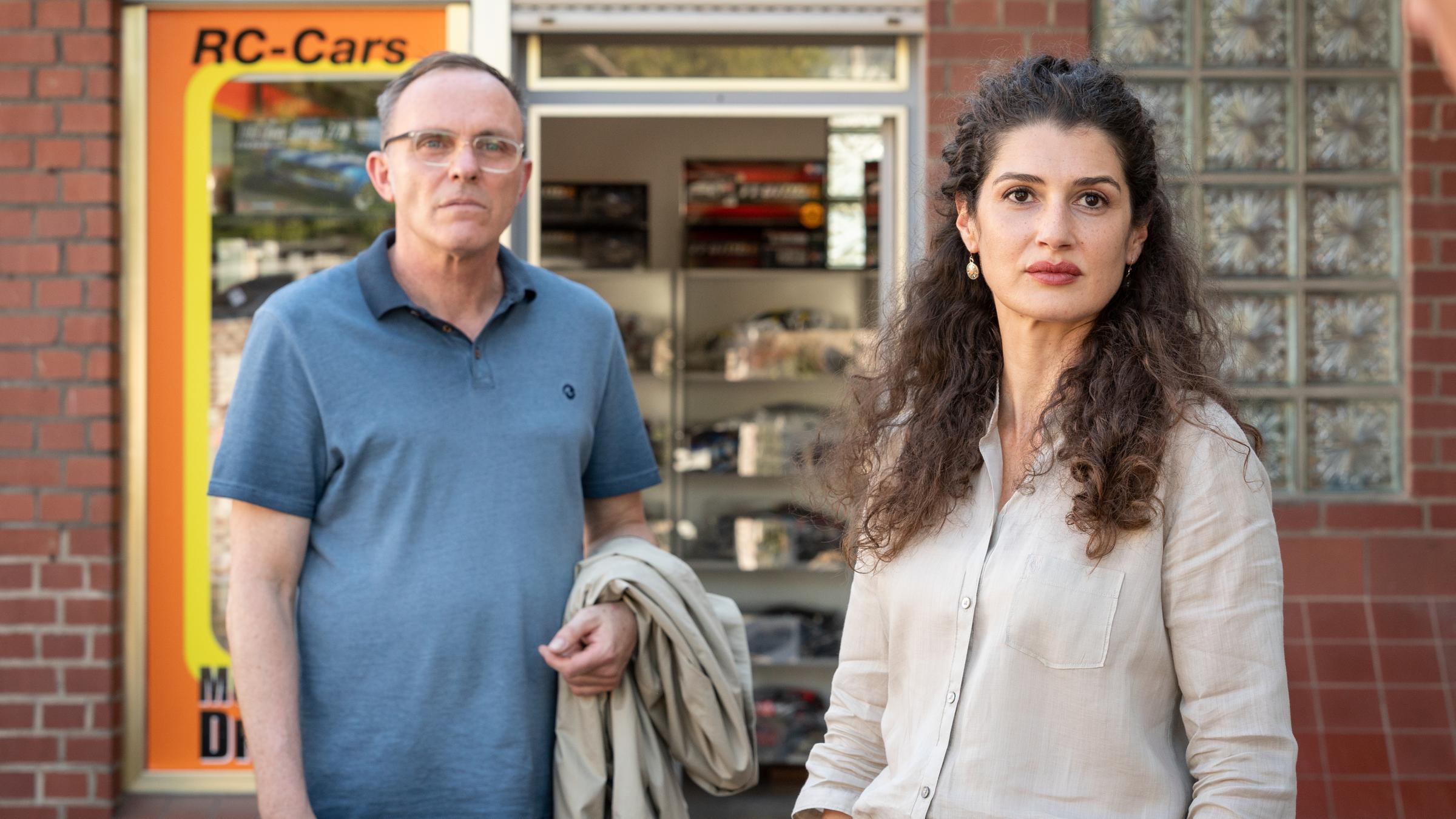 "Wilsberg - Aus heiterem Himmel": Tillmann Drösser (Dirk Martens) und Tahmina Ahmadi (Anastasia Papadopoulou) stehen vor dem Schaufenster eines Modellbaugeschäfts und schauen in die gleiche Richtung.