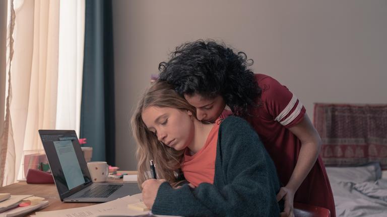 "Loving Her - Loving Franzi": Franzi (Lena Klenke) sitzt am Schreibtisch. Vor ihr steht ein Laptop daneben liegen Papiere. Sie blickt vor sich auf einen Post-it Block, auf dem sie gerade etwas notiert. Hanna (Banafshe Hourmazdi) steht hinter ihr. Sie hat sich zu ihr runtergebeugt und ist im Begriff ihren Nacken zu küssen.