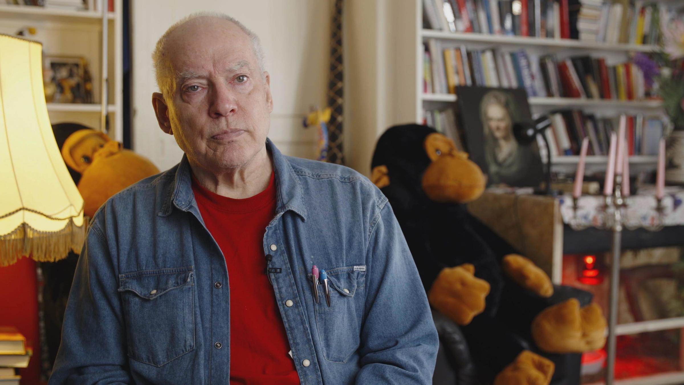 "Queer Cinema": Ein älterer Mann in Jeanshemd und mit kurzen weißen Haaren schaut ernst in die Kamera. Er sitzt vor zwei großen Kuschel-Affen.
