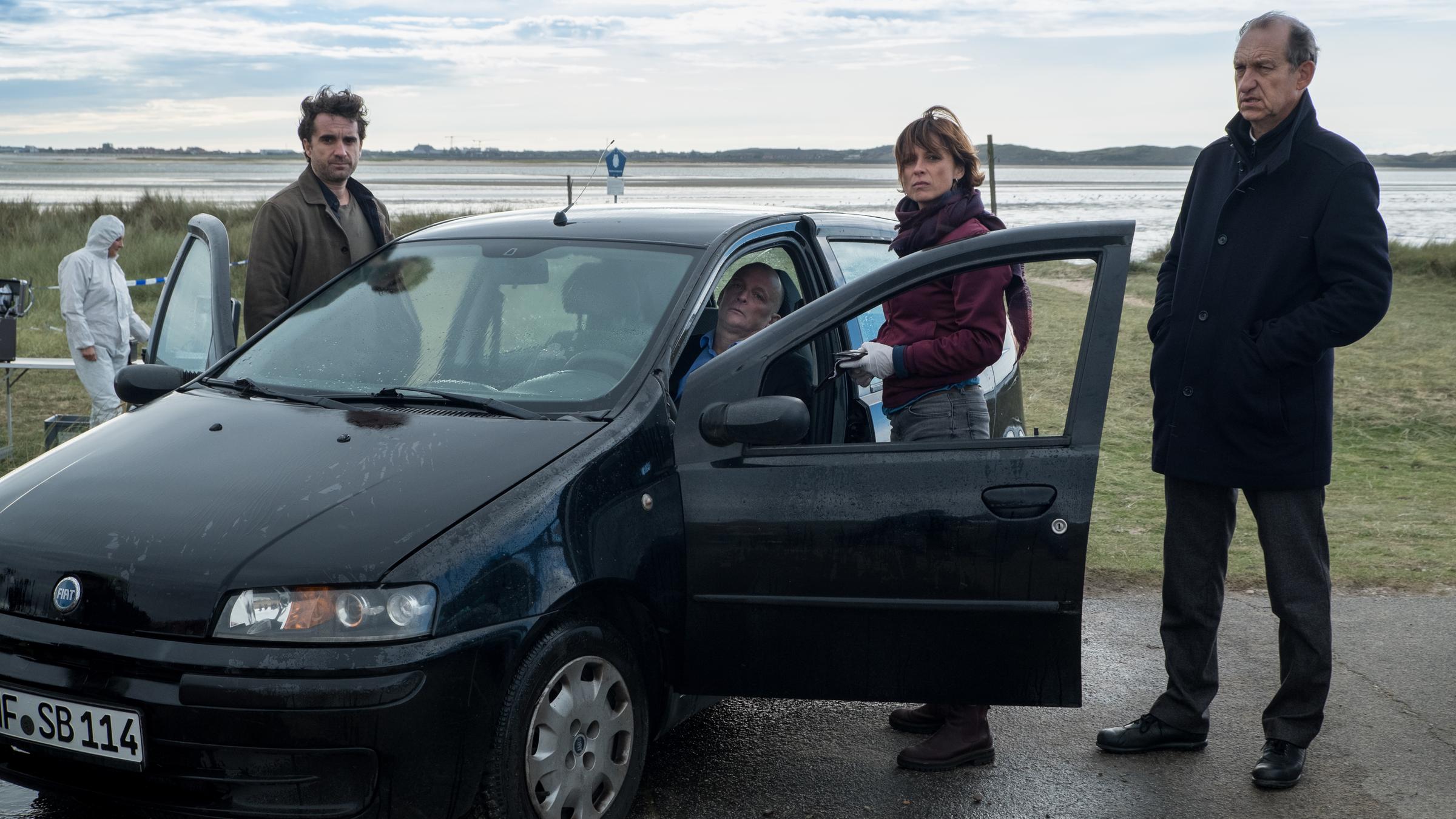 "Nord Nord Mord - Sievers und der schönste Tag": Hinter dem Steuer eines schwarzen Pkw sitzt der tote Hochzeitsvideograf Stefan Borg (Johnny West). Carl Sievers (Peter Heinrich Brix), Ina Behrendsen (Julia Brendler) und Hinnerk Feldmann (Oliver Wnuk ) stehen um das Auto herum. Im Hintergrund sind ein Kollege (Komparse) im weißen Overall und das Wattenmeer zu sehen. Der Himmel ist leicht bewölkt und es ist windig.