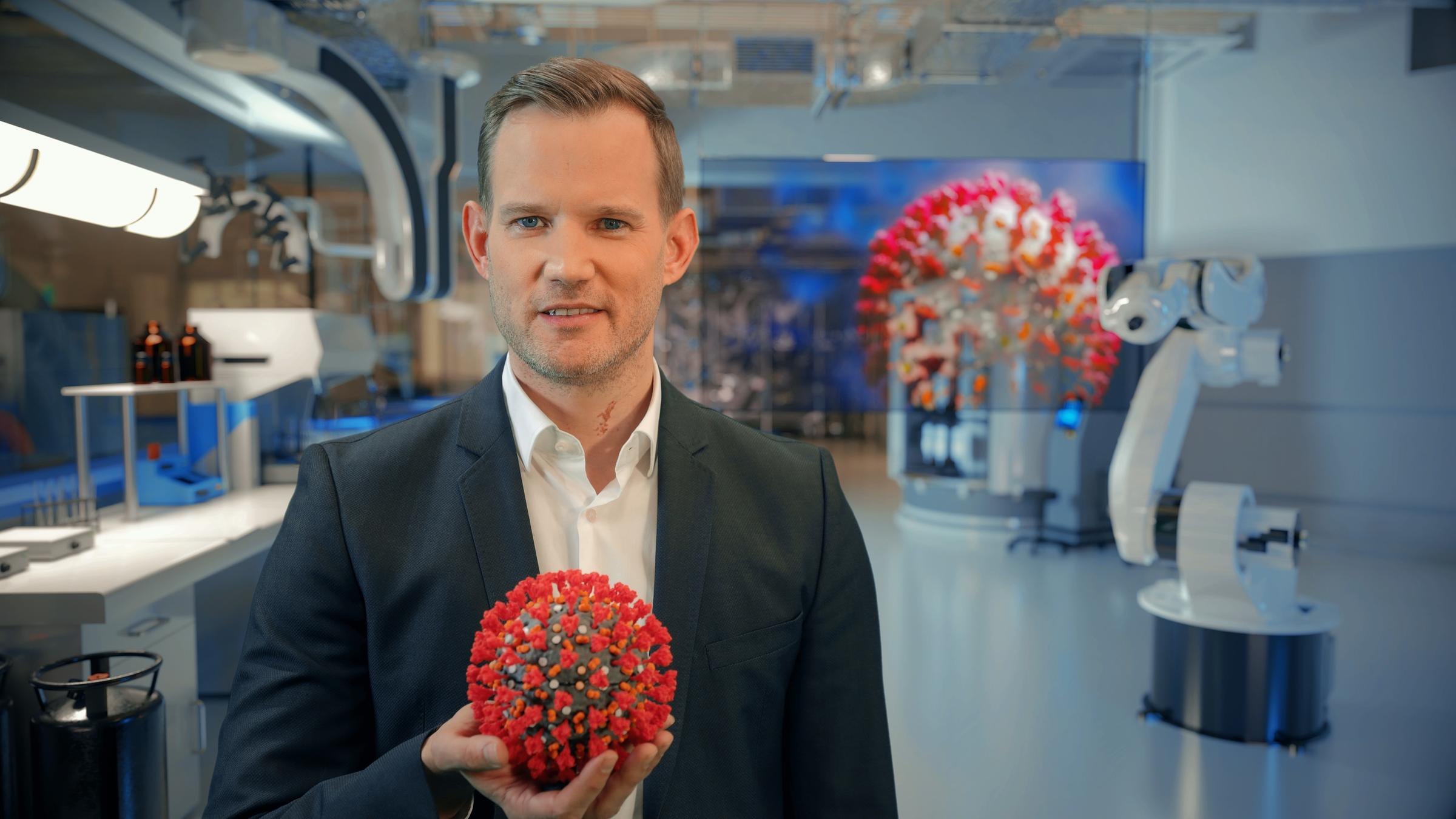 "ZDFzeit: Corona - Pandemie ohne Ende? Fakten mit Hendrik Streeck": Vor einem Labor-Hintergrund steht Hendrik Streeck im anthrazitfarbenem Jackett und hält das Modell eines SARS-CoV-2 mit roten Spikes in der Hand.