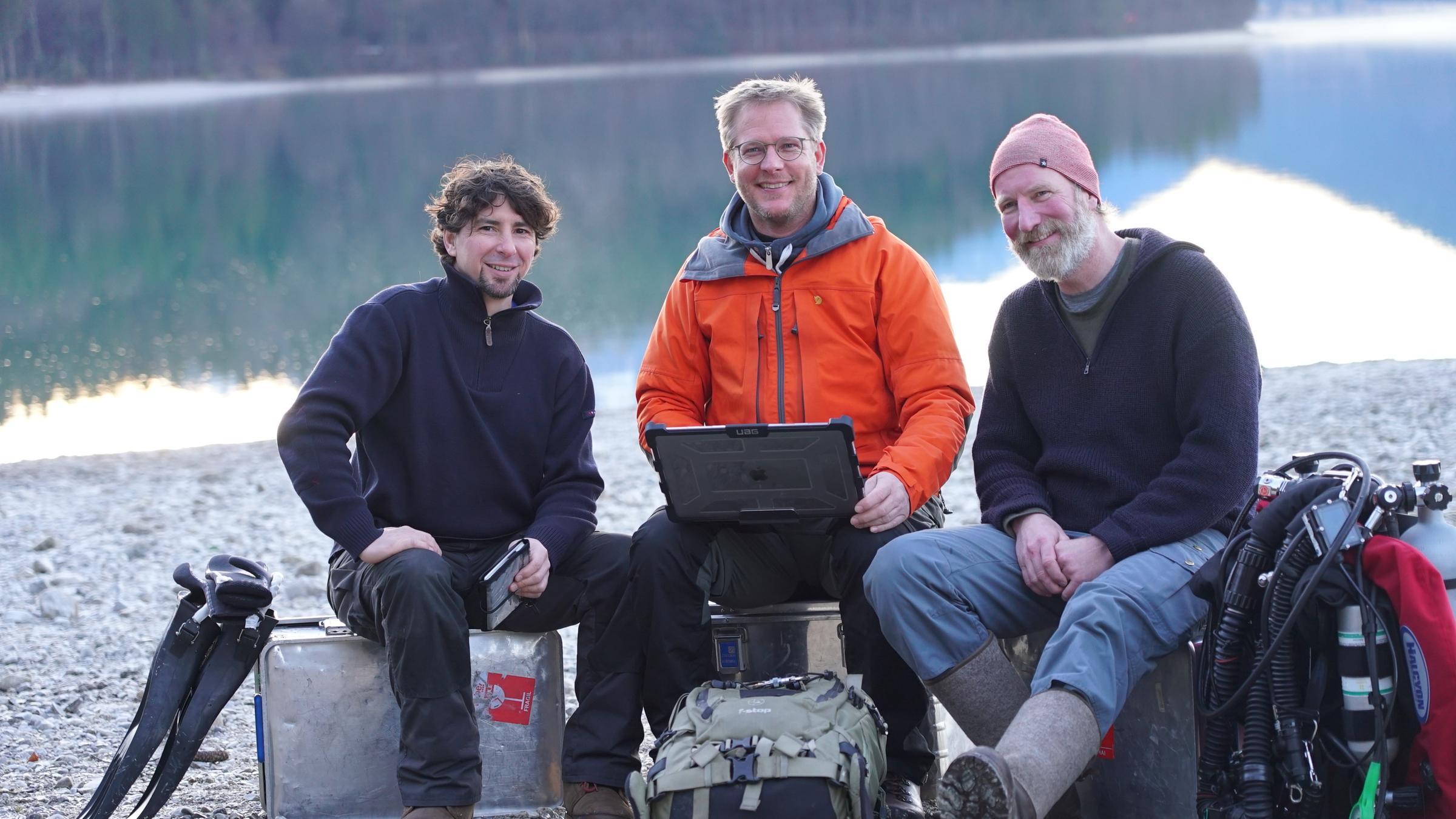 "Terra X: Geheimnisse aus der Tiefe": Dr. Florian Huber, in der Mitte mit roter Jacke, und sein Team, Philipp Schubert und Christian Howe, sitzen vor dem Walchensee.