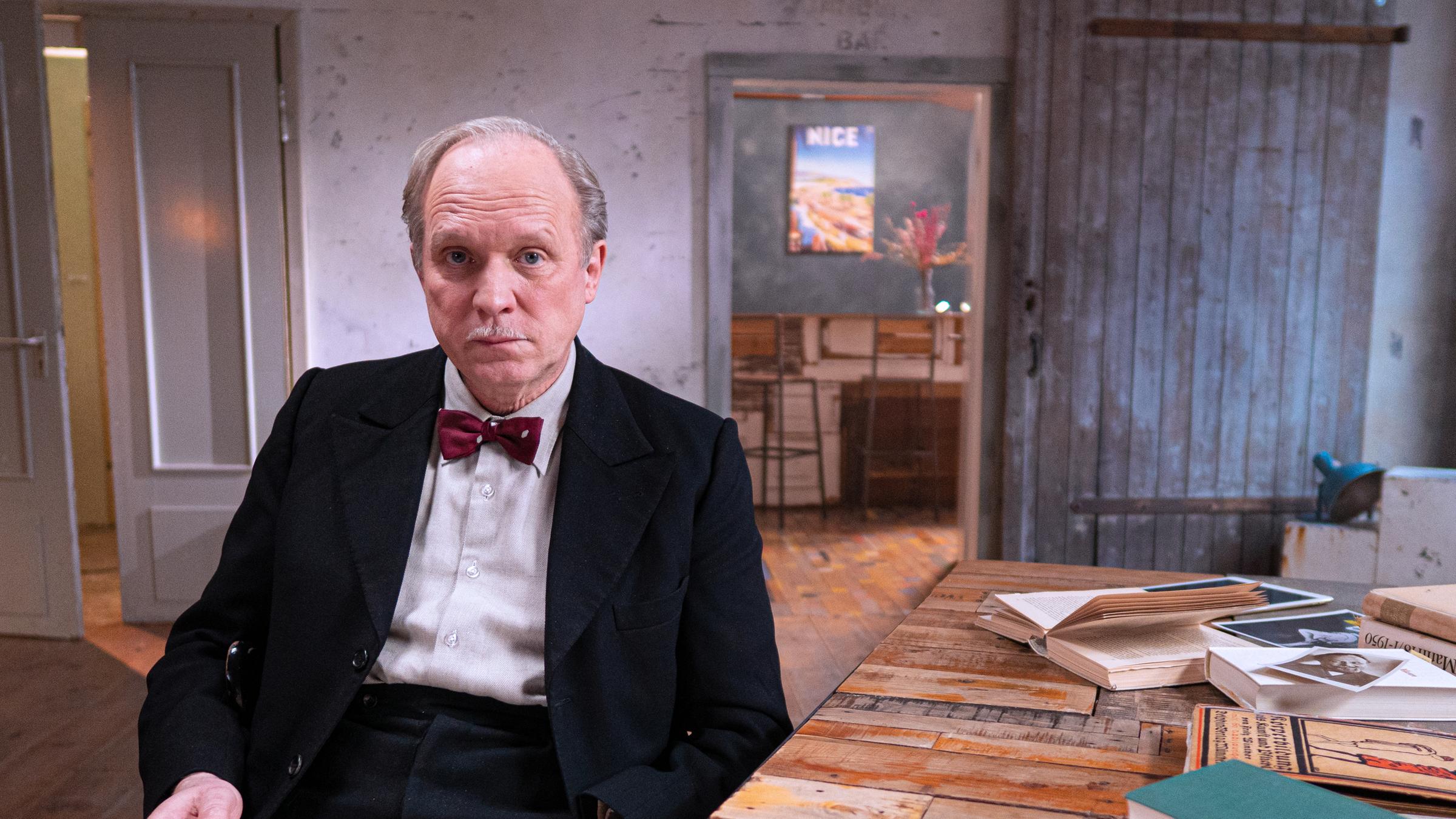  „Heinrich Mann, der unbekannte Rebell:“ Der Schauspieler Ulrich Tukur sitzt an einem Schreibtisch, um ihn herum viele Bücher. Ein Buch hält er in die Hand, er schaut in die Kamera.