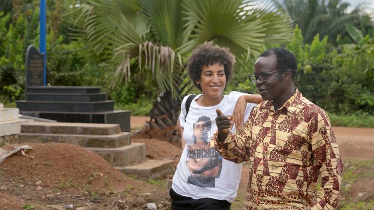 "König Bansah und seine Tochter": Katharina Bansah und ihr Vater Céphas Bansah auf einem Friedhof in Afrika. Im Hintergrund sieht man niedrige Palmen.