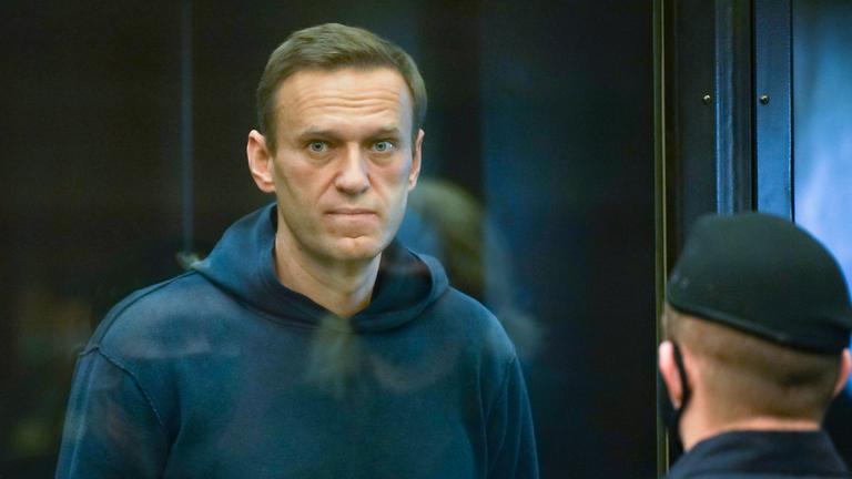 "ZDFzeit: Der Fall Nawalny - Putin, das Gift und die Macht": Alexey Nawalny steht im Käfig im Gericht, bekleidet mit einem dunkelblauen Hoody und schaut direkt durch die Glasscheibe in die Kamera. Mit dem Rücken zur Kamera steht ein Polizist mit Kappe.