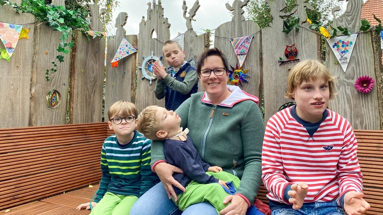 "37° Mama Held - Eine Pflegemutter kämpft für ihre Kinder": Eine Frau mittleren Alters mit kurzem dunklen Haar sitzt inmitten ihrer vier Pflegekinder auf einer Bank im Freien.