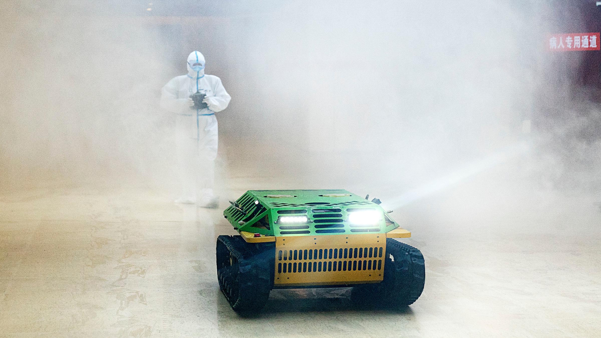 "auslandsjournal - die doku: Der Corona-Kollaps - Wie ein Virus die Welt erschüttert": Ein Seuchenschützer im weißen Schutzanzug läuft hinter einem bunten Roboter-Panzer her, der einen großen desinfiziert. Überall ist weißer Qualm. Der Seuchenschützer steuert den Roboter-Panzer mit einer Fernsteuerung, die er in den Händen hält.