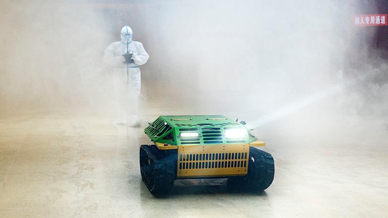 "auslandsjournal - die doku: Der Corona-Kollaps - Wie ein Virus die Welt erschüttert": Ein Seuchenschützer im weißen Schutzanzug läuft hinter einem bunten Roboter-Panzer her, der einen großen desinfiziert. Überall ist weißer Qualm. Der Seuchenschützer steuert den Roboter-Panzer mit einer Fernsteuerung, die er in den Händen hält.