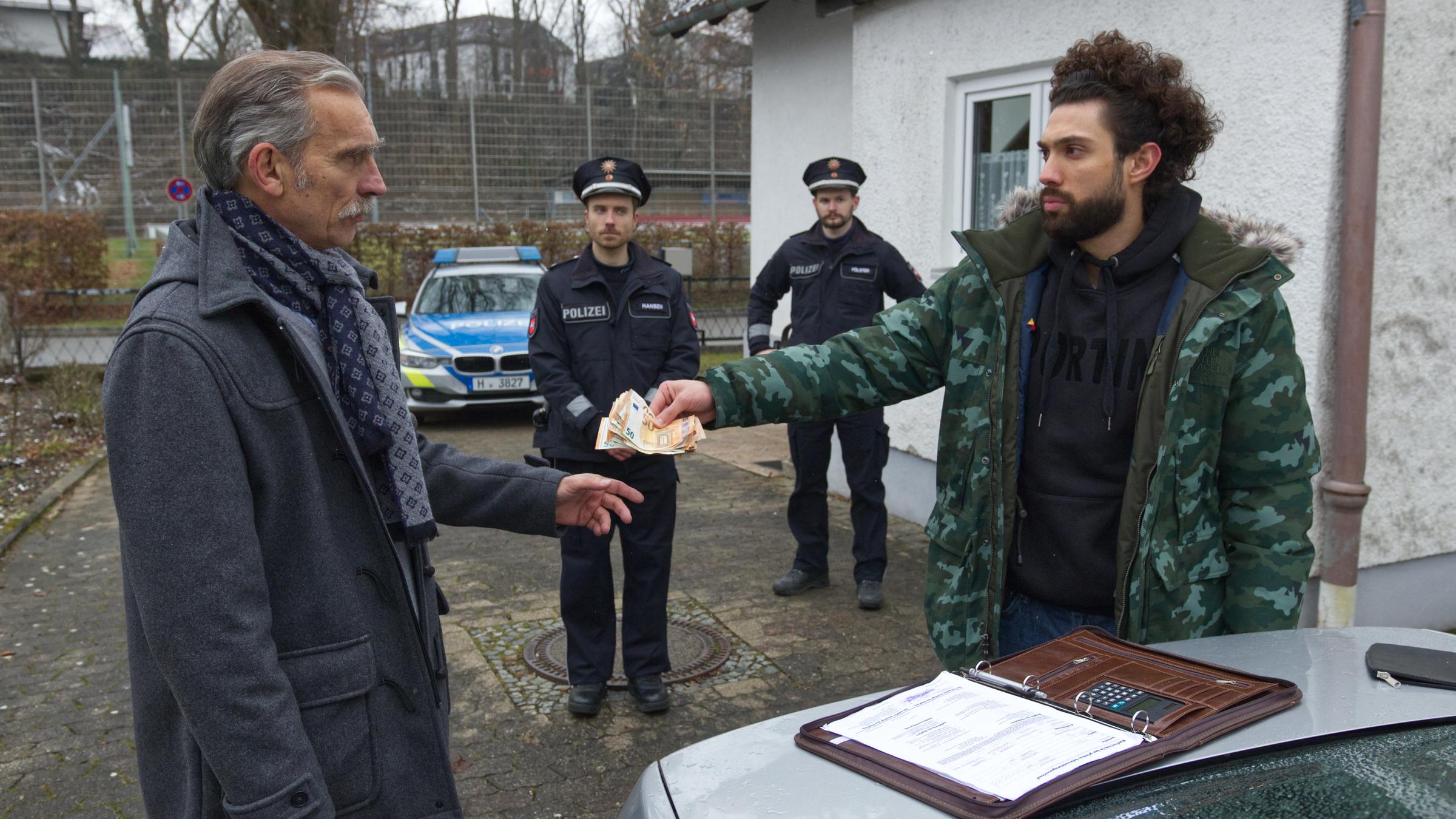 "Aktenzeichen XY-Spezial: Vorsicht, Betrug!": Vor einem Haus übergibt ein junger Mann einem älteren Herrn mehrere Geldscheine. Zwei Polizeibeamte stehen im Hintergrund und beobachten das Geschehen.