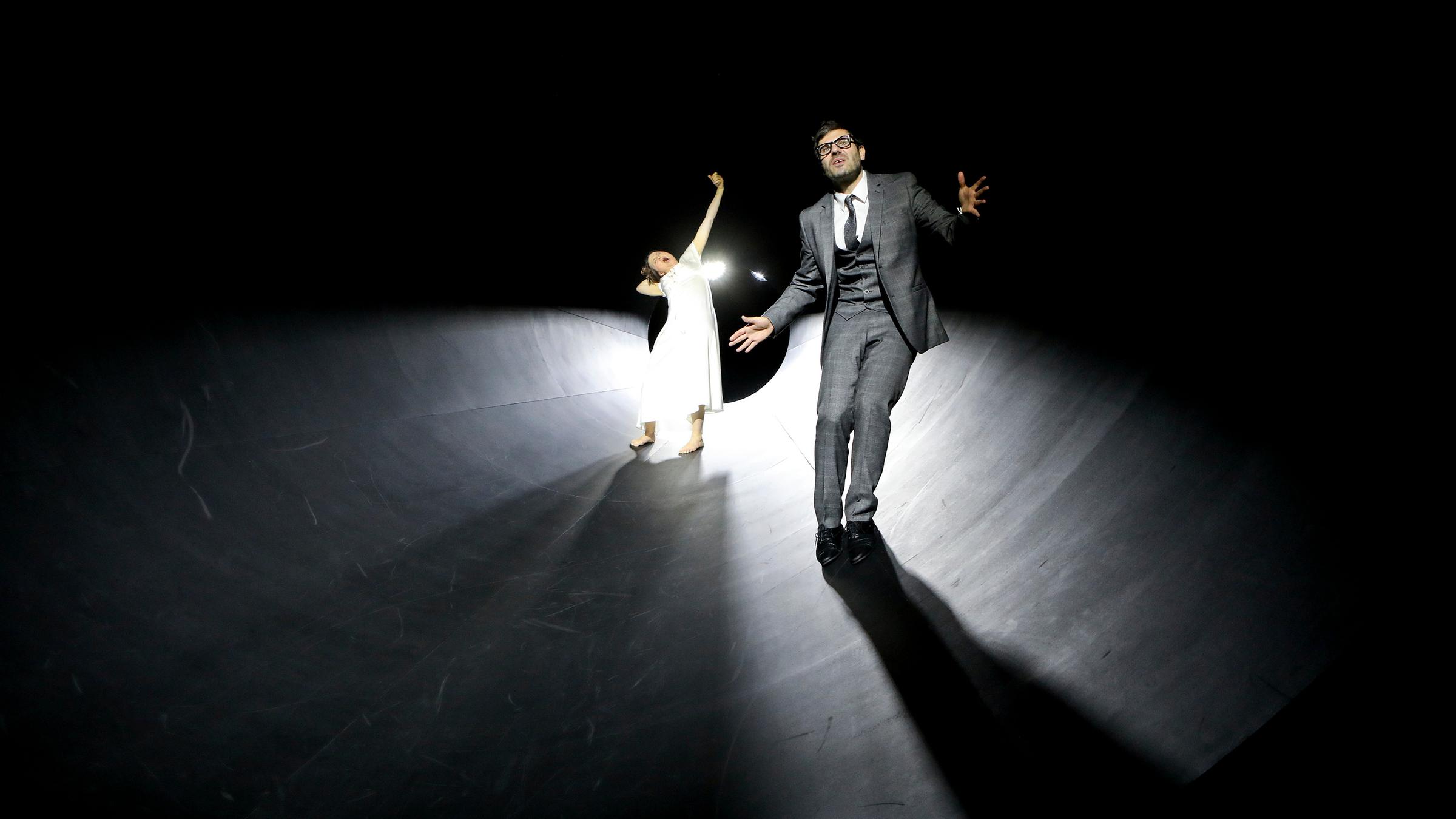 "Starke Stücke: Graf Öderland": In einem schwarzen Trichter, der von hinten beleuchtet wird, befinden sich zwei Personen. Im Hintergrund eine Frau in einem weißen Kleid, die mit erhobenem linkem Arm und nach links geneigtem Kopf das eigene Erhängen simuliert. Im Vordergrund rechts, mit dem Rücken zu ihr, ein Mann mit dunklen, kurzen Haaren und Brille. Er hat die Arme leicht ausgebreitet und trägt einen dreiteiligen grauen Anzug.