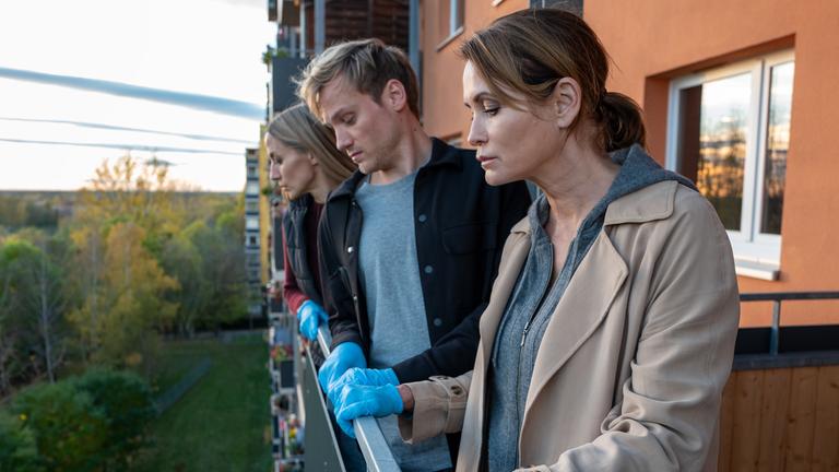 "Das Quartett - Die Tote vom Balkon": Maike (Anja Kling), Linus (Anton Spieker) und Pia (Annika Blendl) stehen nebeneinander auf einem Balkon und blicken in die Tiefe. Im Hintergrund sind weitere Balkone des Hauses zu erkennen.