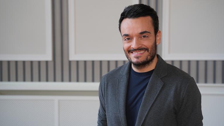 "Heimliche Helden": Giovanni Zarrella steht vor einer gestreiften Wand und lächelt, gekleidet in einem grauen Jackett.