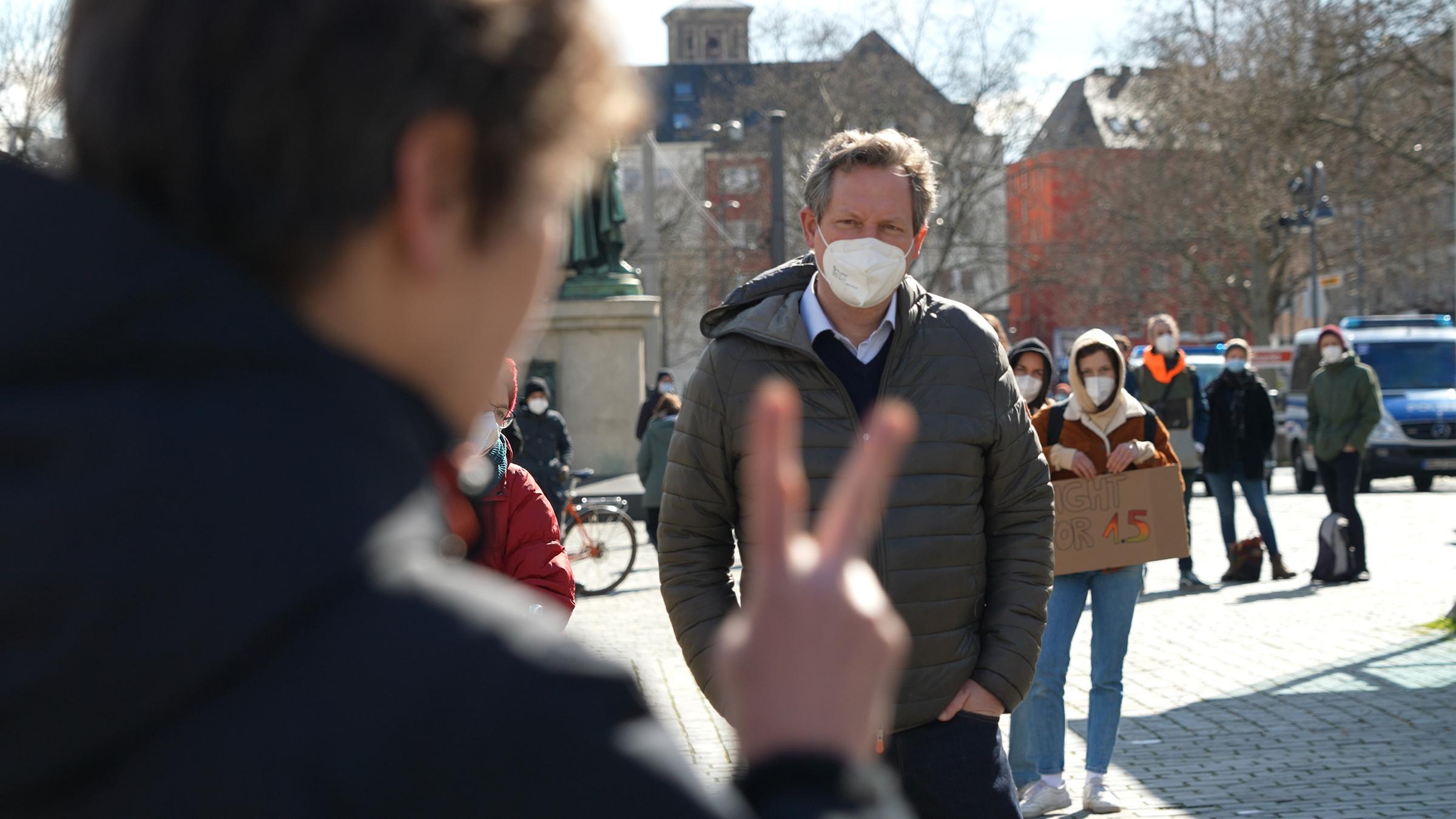 "Die sieben Bösen - Eckart von Hirschhausen: Der Sünde auf der Spur": Eckart von Hirschhausen steht auf einem Platz vor einer Menschengruppe. Er trägt einen Mund-Nasen-Schutz und schaut zu einem Mann, der mit dem Rücken zur Kamera steht und die Finger seiner rechten Hand zum Friedenszeichen erhoben hat.