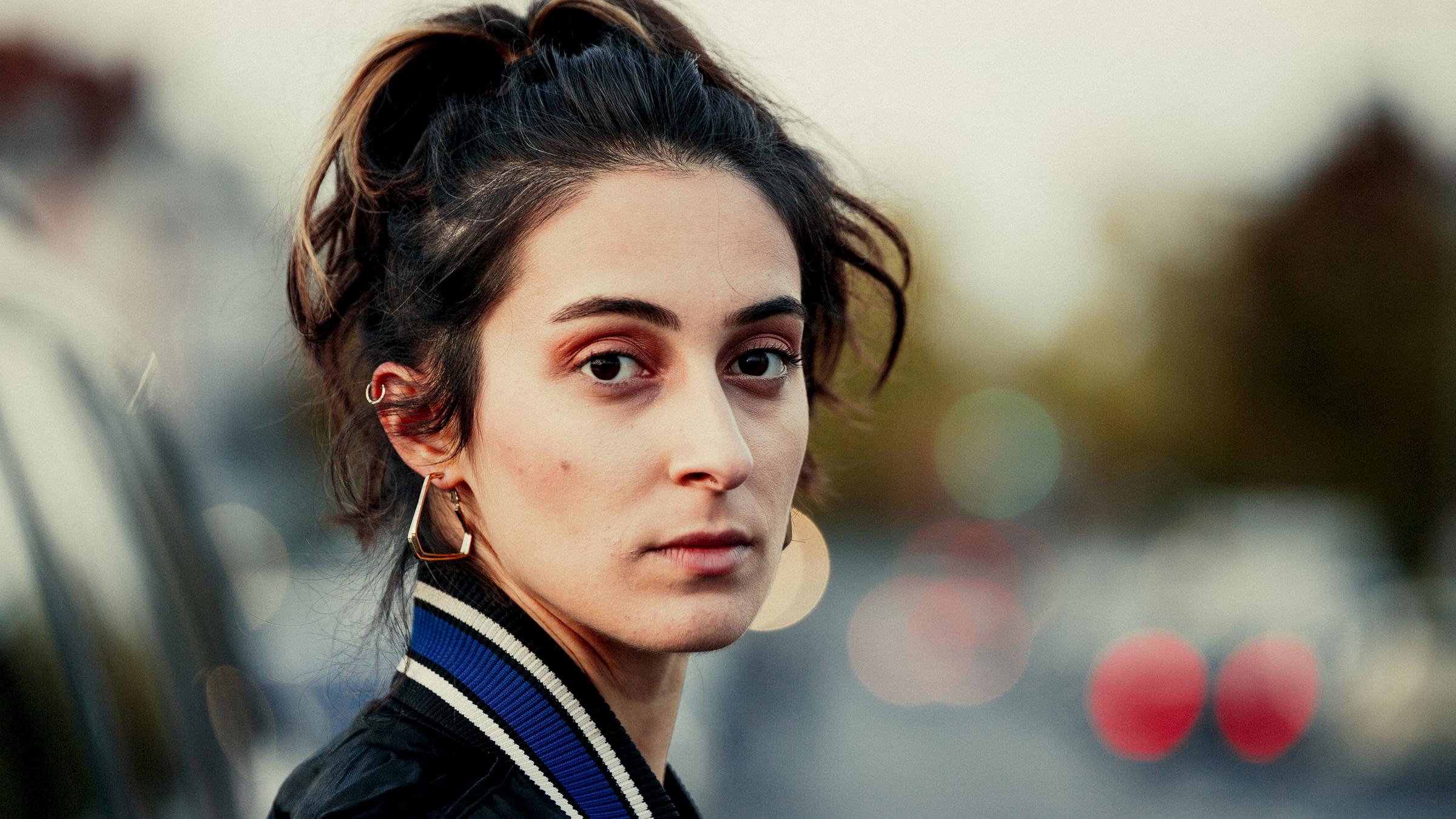 "Ansonsten geht's mir gut": Porträtaufnahme einer jungen Frau, die in die Kamera blickt, im Hintergrund verschwommen die Rücklichter von Autos.