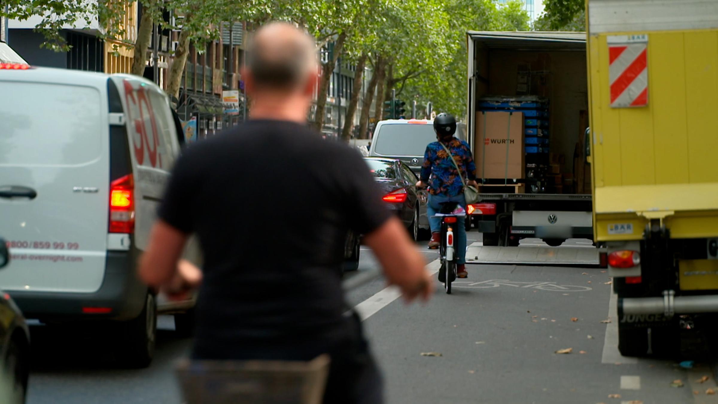 "Hallo Deutschland hautnah: Der neue Fahrradboom - Lust und Frust im Großstadtdschungel": Verkehrsreiche Innenstadt-Straße. Pkw, Lieferfahrzeuge und Radfahrer drängen sich dicht aneinander vorbei. Der Radweg ist durch einen Sprinter blockiert.