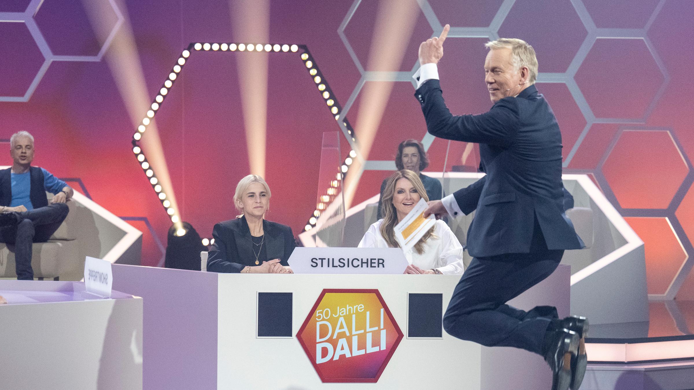 "50 Jahre Dalli Dalli - die große Jubiläumsshow": Caro Daur und Frauke Ludowig sitzen - durch eine Plexiglaswand getrennt - hinter einem Ratepult. Sie lachen und schauen Johannes  B. Kerner an, der vor ihnen hochspringt und in die Kamera lacht. im Hintergrund sind Michael Mittermeier und Adele Neuhauser zu sehen.