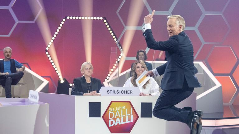 "50 Jahre Dalli Dalli - die große Jubiläumsshow": Caro Daur und Frauke Ludowig sitzen - durch eine Plexiglaswand getrennt - hinter einem Ratepult. Sie lachen und schauen Johannes  B. Kerner an, der vor ihnen hochspringt und in die Kamera lacht. im Hintergrund sind Michael Mittermeier und Adele Neuhauser zu sehen.