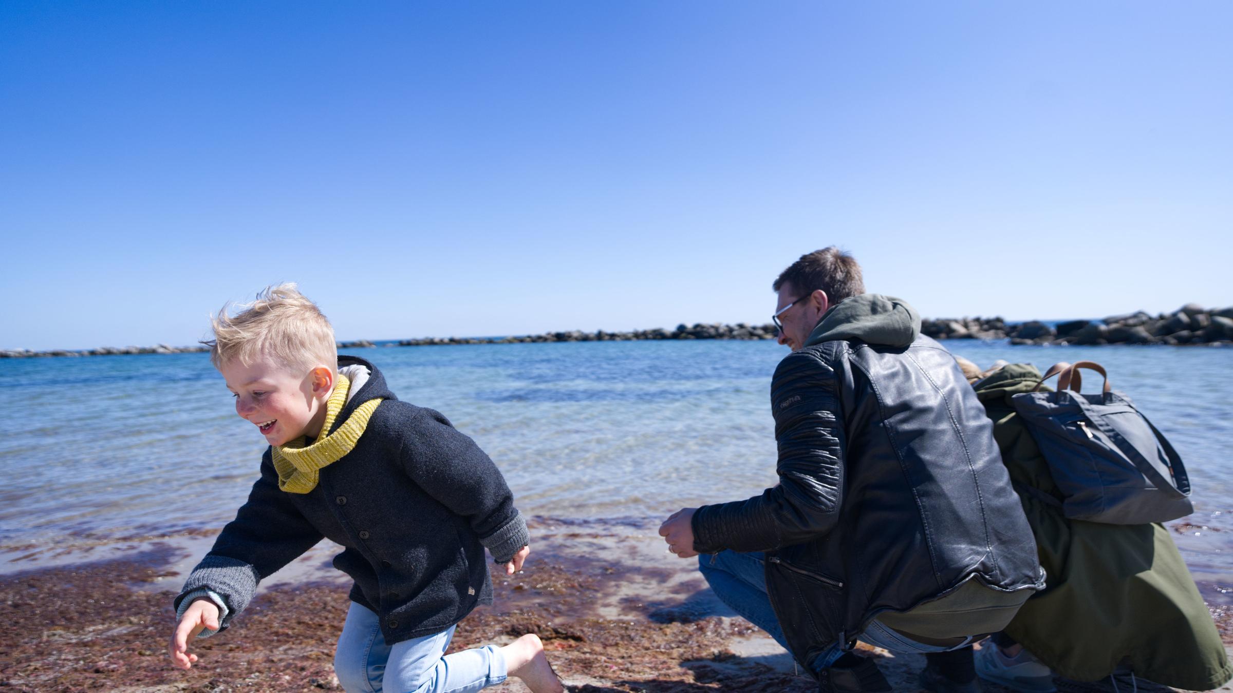 "Wachgeküsst - Urlaub mal wieder in Deutschland": Eine Familie sitzt am Ostseestrand und blickt auf das Meer. Ihr kleiner Sohn läuft lachend nach links. Der Himmel ist blau und im Hintergrund sieht man einen bewaldeten Küstensaum.