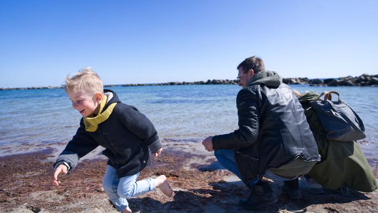 "Wachgeküsst - Urlaub mal wieder in Deutschland": Eine Familie sitzt am Ostseestrand und blickt auf das Meer. Ihr kleiner Sohn läuft lachend nach links. Der Himmel ist blau und im Hintergrund sieht man einen bewaldeten Küstensaum.