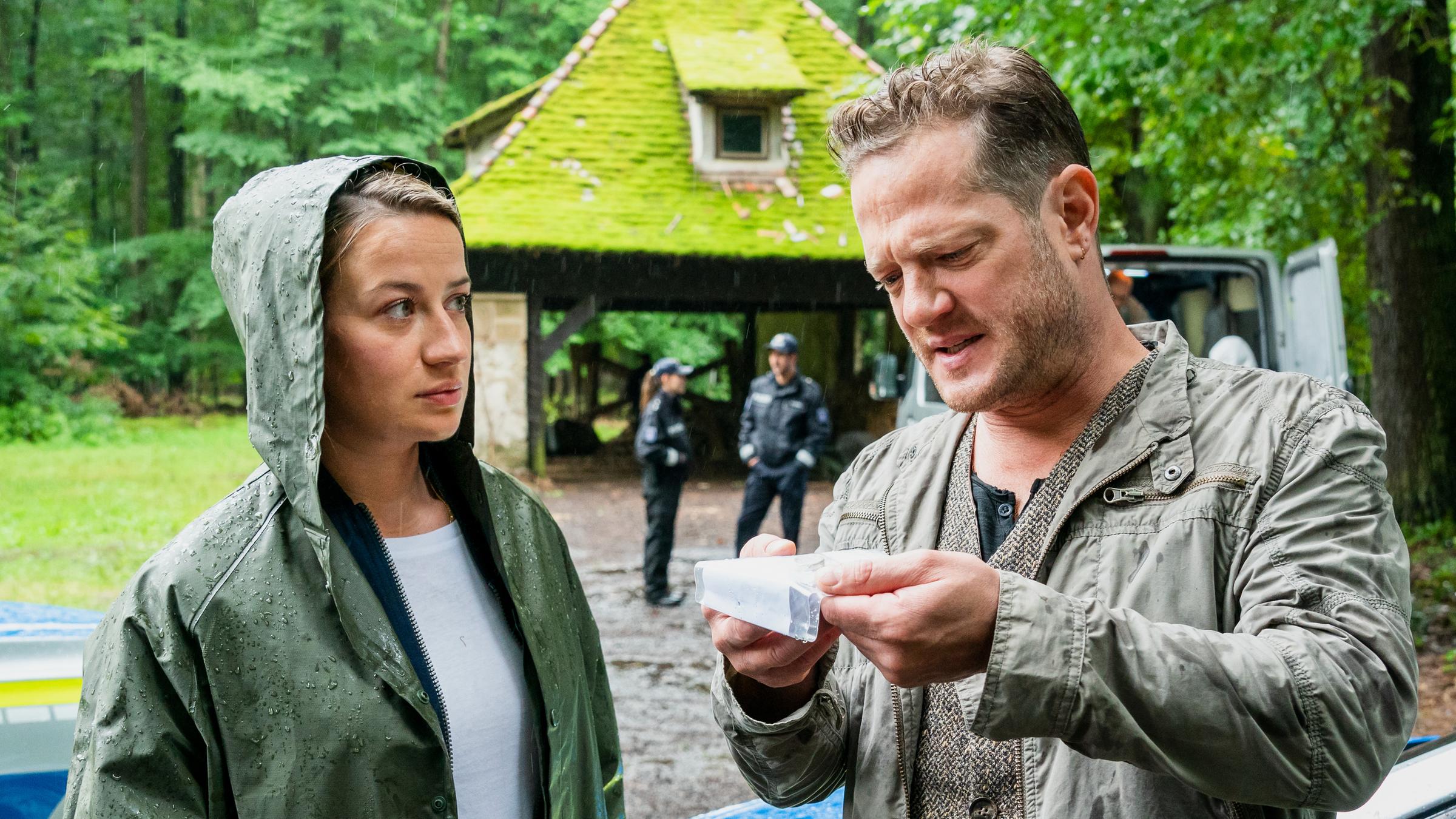 "Theresa Wolff - Waidwund": Theresa Wolff (Nina Gummich) hat ihre Regenjacke an und blickt Bruno Lewandowski (Aurel Manthei) an, während dieser mit zusammengekniffenen Augen eine kleine Plastiktüte inspiziert. Im Hintergrund sieht man einen Unterstand im Wald, vor dem zwei uniformierte Polizeibeamte stehen.
