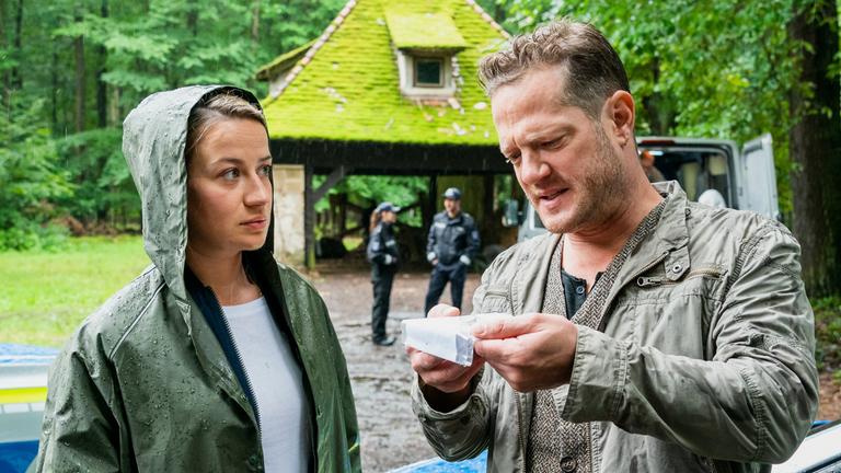 "Theresa Wolff - Waidwund": Theresa Wolff (Nina Gummich) hat ihre Regenjacke an und blickt Bruno Lewandowski (Aurel Manthei) an, während dieser mit zusammengekniffenen Augen eine kleine Plastiktüte inspiziert. Im Hintergrund sieht man einen Unterstand im Wald, vor dem zwei uniformierte Polizeibeamte stehen.