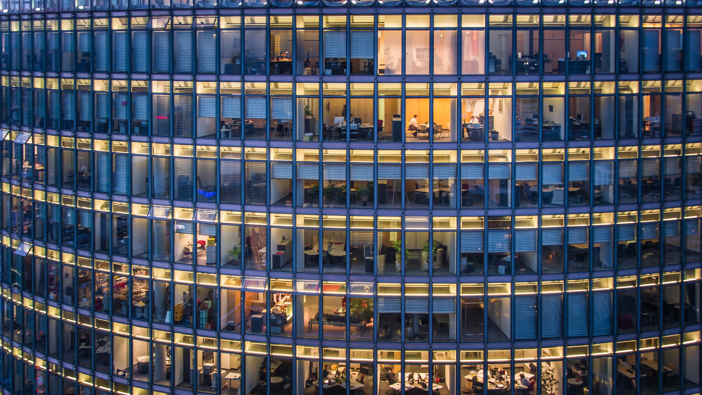 "Big Four - Wirtschaftsprüfer unter Verdacht": Hell erleuchtete Büros in einem Hochhaus. Der Blick durch die Glasfassade zeigt verschiedene Arbeitsplätze, vereinzelt mit Personen am Schreibtisch sitzend.