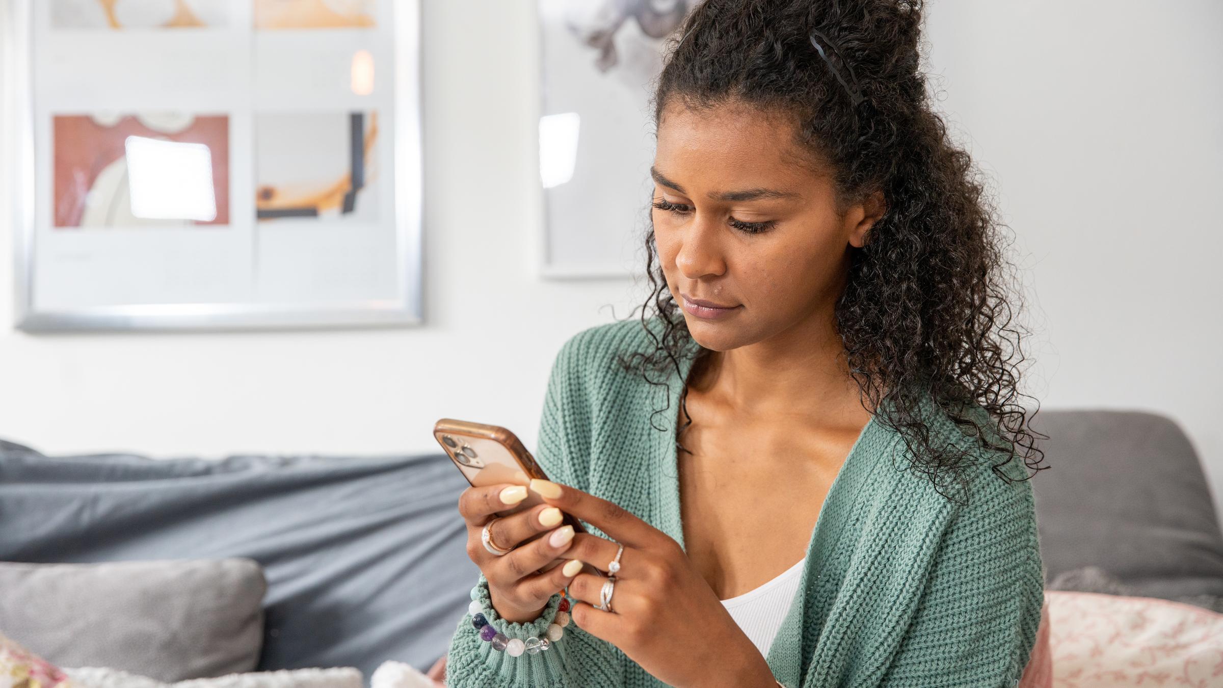 "37° Cybermobbing - Angriff aus dem Netz": Lijana Kaggwa, eine junge Frau mit schwarzen, gelockten Haaren sitzt auf einem Stuhl und hält ein Handy in beiden Händen. Im Hintergrund ist eine Couch zu sehen.