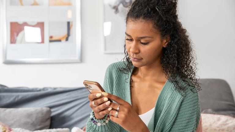 "37° Cybermobbing - Angriff aus dem Netz": Lijana Kaggwa, eine junge Frau mit schwarzen, gelockten Haaren sitzt auf einem Stuhl und hält ein Handy in beiden Händen. Im Hintergrund ist eine Couch zu sehen.