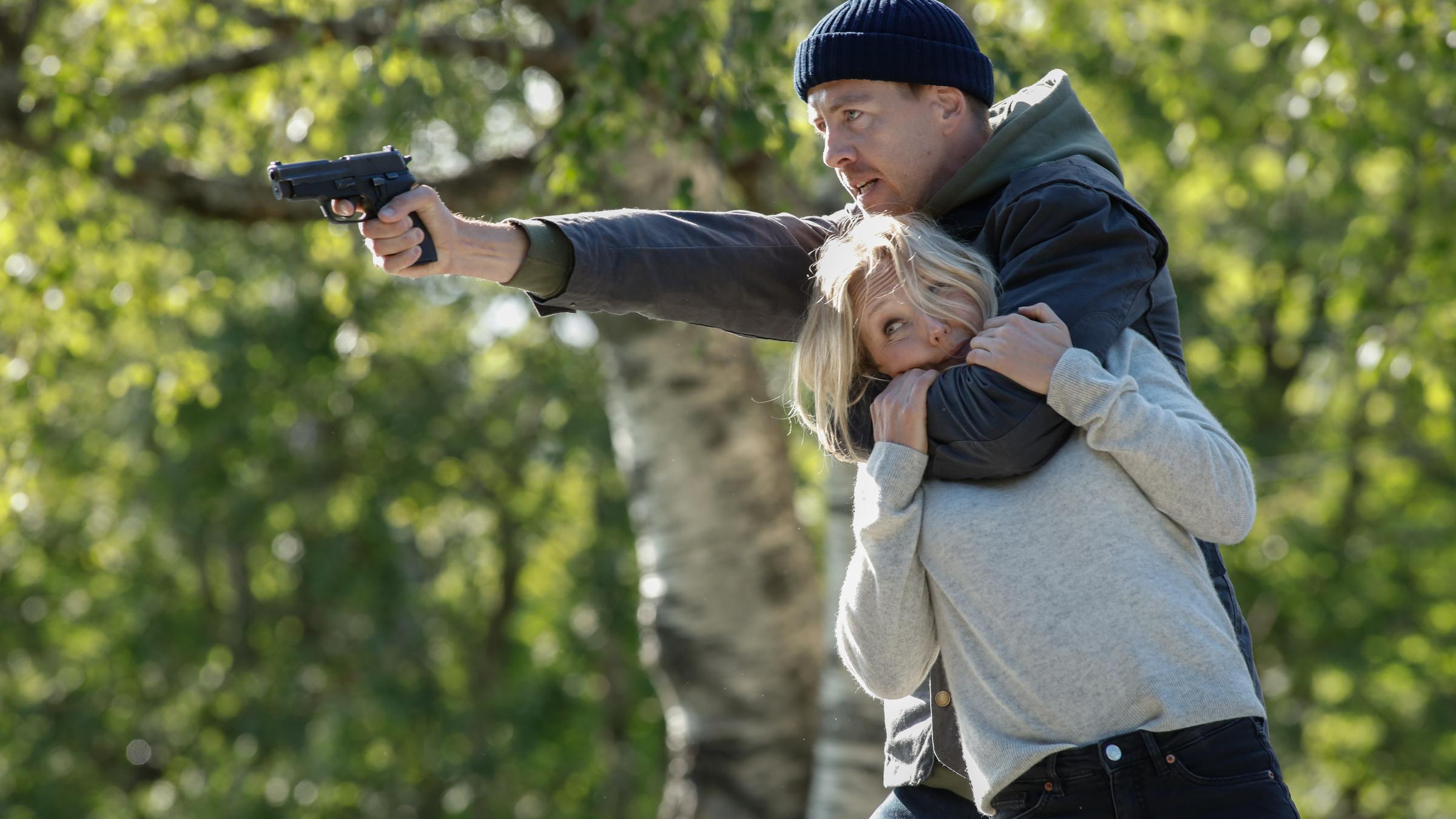 "Furia (1)": Im Wald hält Asgeir (Pål Sverre Hagen) mit seinem linken Arm Ragna (Ine Marie Wilmann) im Schwitzkasten, während er mit der Pistole in seiner rechten Hand auf jemanden außerhalb des Bildes zielt.