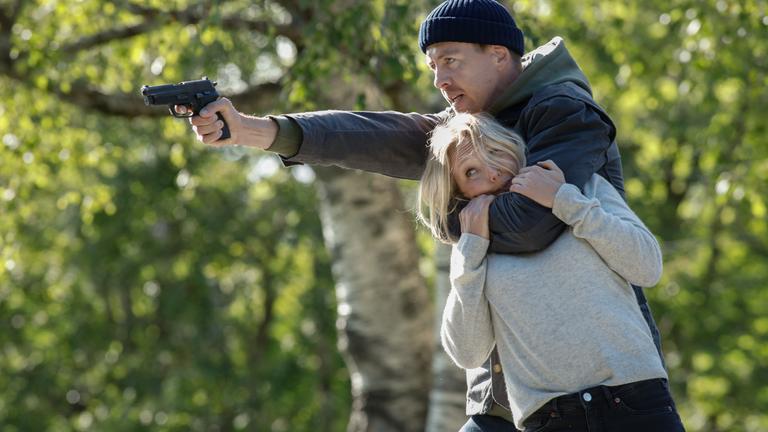 "Furia (1)": Im Wald hält Asgeir (Pål Sverre Hagen) mit seinem linken Arm Ragna (Ine Marie Wilmann) im Schwitzkasten, während er mit der Pistole in seiner rechten Hand auf jemanden außerhalb des Bildes zielt.