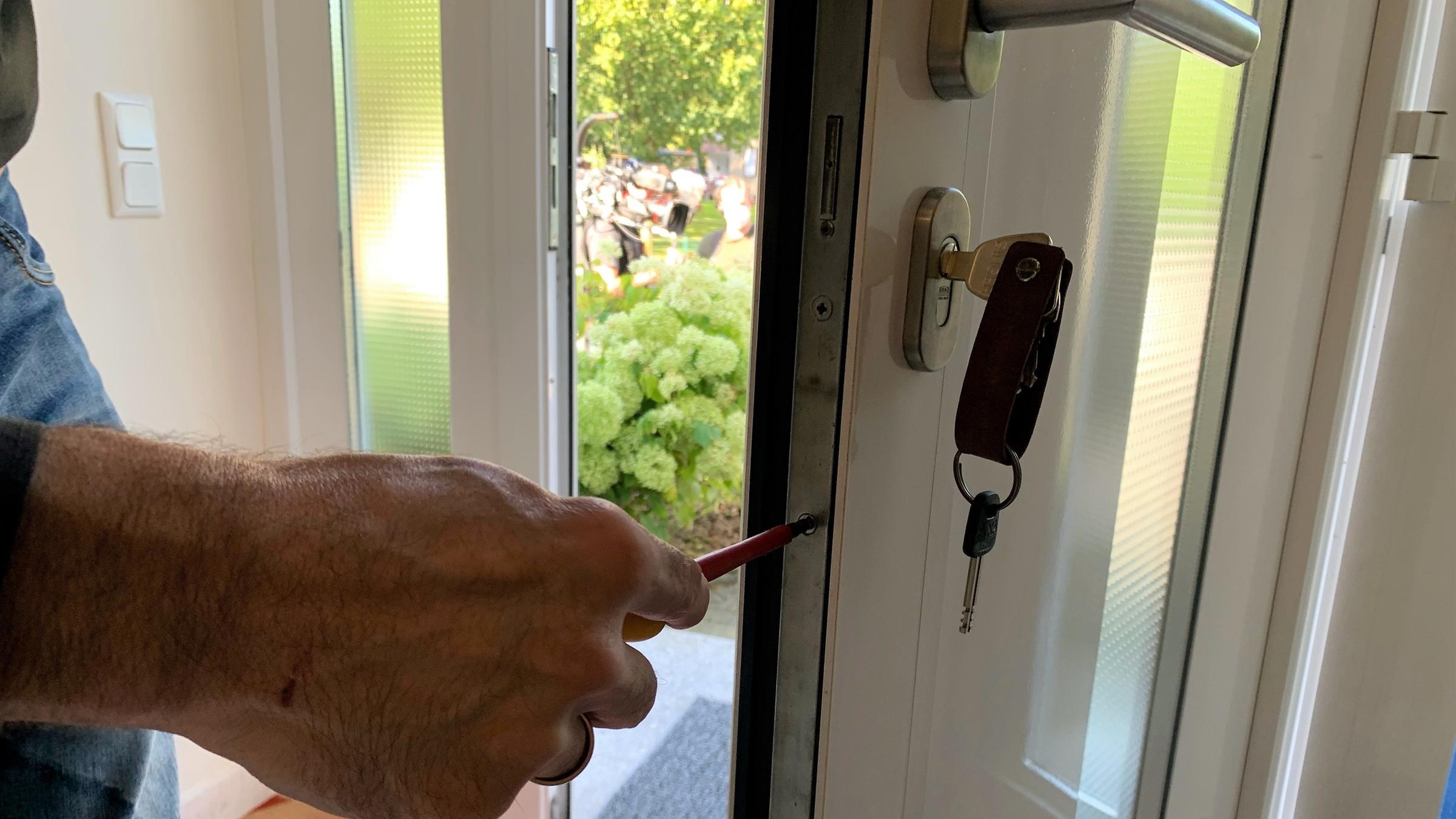 "ZDF.reportage: Albtraum Einbruch": Im rechten Vordergrund des Bildes ist eine halb geöffnete Tür zu erkennen. In der Innenseite der Tür steckt ein Schlüssel. Im linken Vordergrund ist eine Hand zu sehen. Diese hält einen Schraubenzieher in der Hand, der in die Innenseite der geöffneten Tür eingeführt wird. Ein neues Türschloss wird verbaut. Es handelt sich um eine Haustür. Diese ist weiß und mit Milchglas besetzt. Hinter der geöffneten Tür sind Büsche zu erkennen. Auf dem Boden sieht man einen rot-orangenen Teppich.