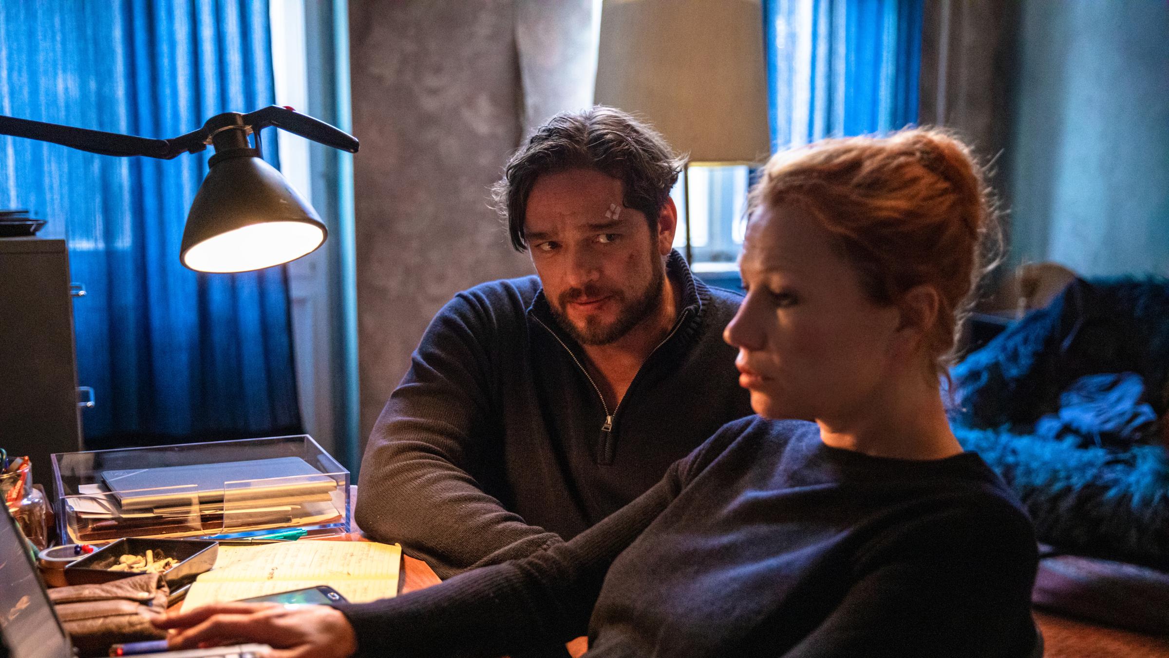 "Dengler - Kreuzberg Blues": Olga Illiescu (Birgit Minichmayr) sitzt daheim an ihrem Schreibtisch, der vollgestellt ist mit Unterlagen und Technik. Sie tippt gerade etwas auf ihrem Laptop, der direkt vor ihr steht. Dengler (Ronald Zehrfeld) sitzt neben ihr, ist ihr mit dem ganzen Körper zugewandt und blickt sie kritisch an.